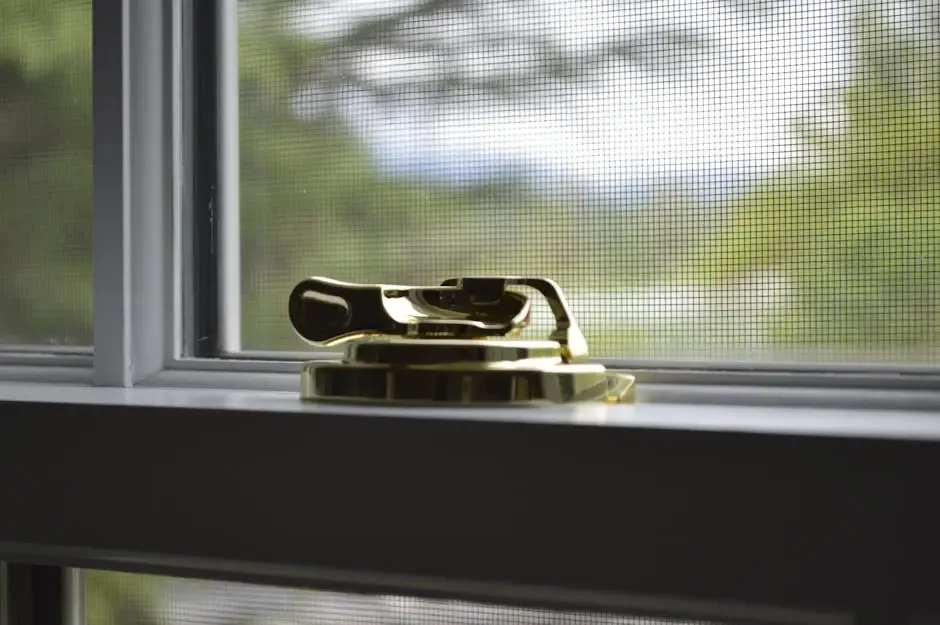 A close-up of a shiny gold window lock resting on a white windowsill, with a mesh screen and blurred greenery visible through the window.