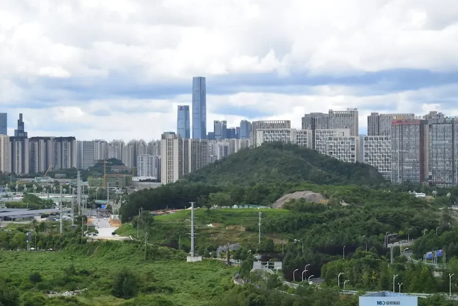 A panoramic view of a modern cityscape featuring tall skyscrapers, including a notable glass building, set against a backdrop of a green hill and scattered residential buildings, under a partly cloudy sky.